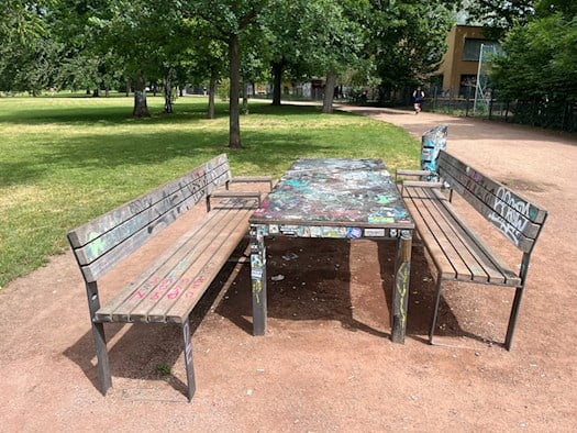 Eine hölzerne, barrierefreie Sitzgruppe mit graffiti-verzierter Tischplatte und zwei Holzbänken auf dem Alaunplatz in Dresden Neustadt.