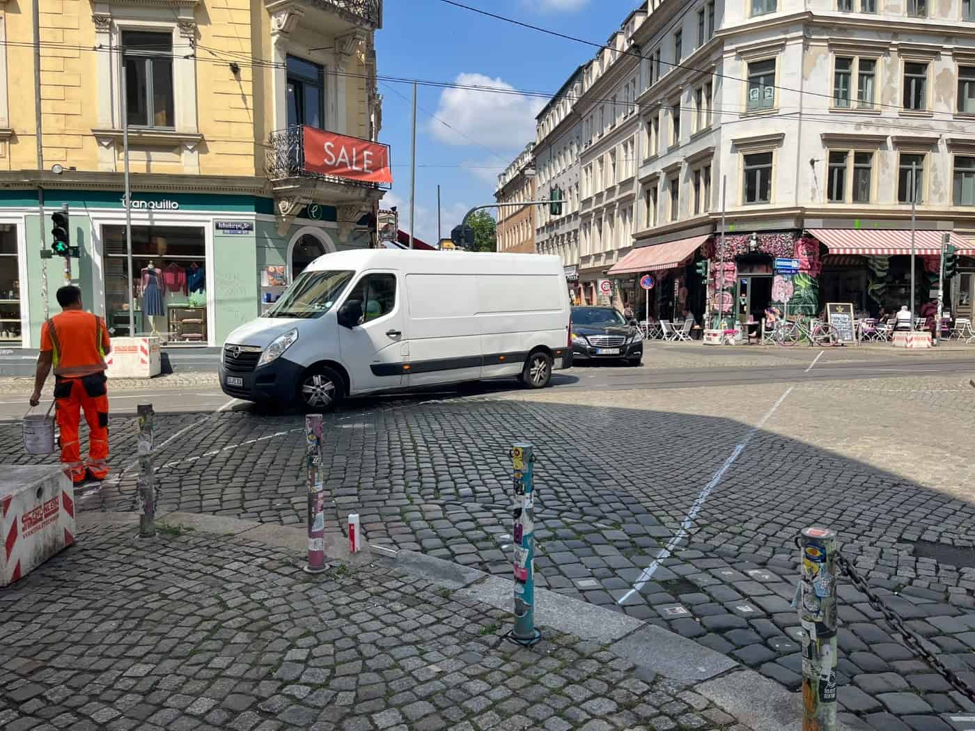 Kopfsteinpflaster-Kreuzung mit abbiegendem weißem Lieferwagen. Links steht ein Bauarbeiter in orangefarbener Warnkleidung mit Farbeimer neben frisch gezogenen weißen Linien. Im Hintergrund Eckhäuser des Szeneviertels.
