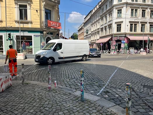 Kopfsteinpflaster-Kreuzung mit abbiegendem weißem Lieferwagen. Links steht ein Bauarbeiter in orangefarbener Warnkleidung mit Farbeimer neben frisch gezogenen weißen Linien. Im Hintergrund Eckhäuser des Szeneviertels.