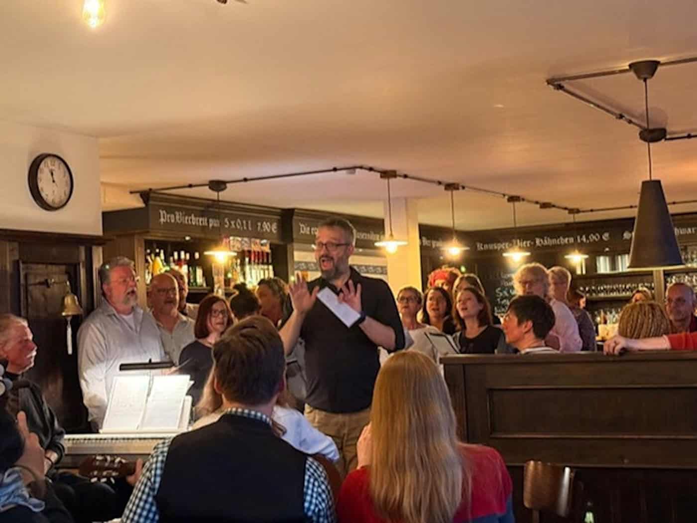 Foto: in einer Kneipe singen zwei Dutzend Erwachsene. Im Vordergrund spielt jemand Keyboard mit Noten. Im Hintergrund stehen Regale mit Flaschen und Gläsern.