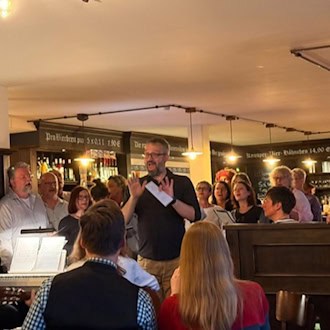 Foto: in einer Kneipe singen zwei Dutzend Erwachsene. Im Vordergrund spielt jemand Keyboard mit Noten. Im Hintergrund stehen Regale mit Flaschen und Gläsern.