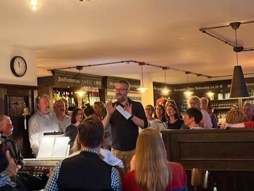 Foto: in einer Kneipe singen zwei Dutzend Erwachsene. Im Vordergrund spielt jemand Keyboard mit Noten. Im Hintergrund stehen Regale mit Flaschen und Gläsern.