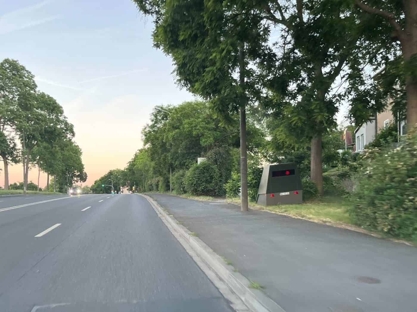 Ein olivgrün lackierter, kantiger Blitzer-Anhänger (Enforcement Trailer) steht am Straßenrand der Meißner Landstraße im Gras vor Bäumen und Hecken. Auf der robusten Außenhülle sind leichte Spuren des gelöschten Brandes zu sehen.