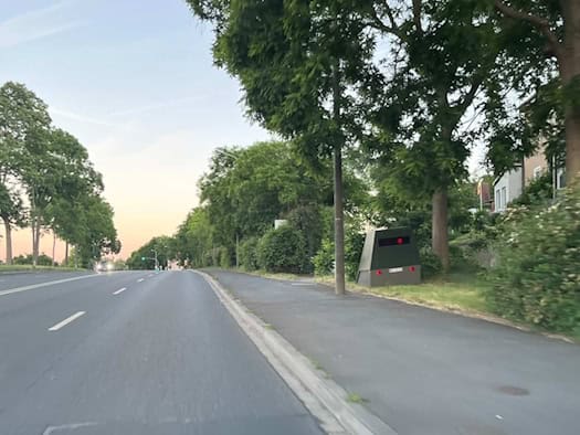 Ein olivgrün lackierter, kantiger Blitzer-Anhänger (Enforcement Trailer) steht am Straßenrand der Meißner Landstraße im Gras vor Bäumen und Hecken. Auf der robusten Außenhülle sind leichte Spuren des gelöschten Brandes zu sehen.