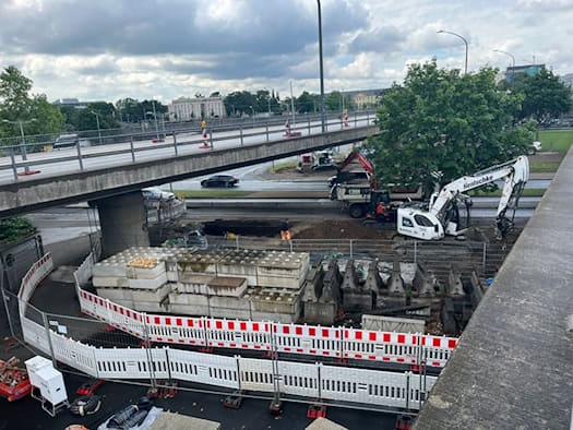 Blick von oben auf eine mit rot-weißen Bauzäunen abgesperrte Baustelle direkt unter einer mehrspurigen Betonbrücke. Ein großer weißer Kettenbagger mit der Aufschrift „Hentschke“ sowie ein kleinerer Bagger stehen auf aufgewühlter Erde neben gestapelten Betonblöcken. Auf der Brücke im Hintergrund flie