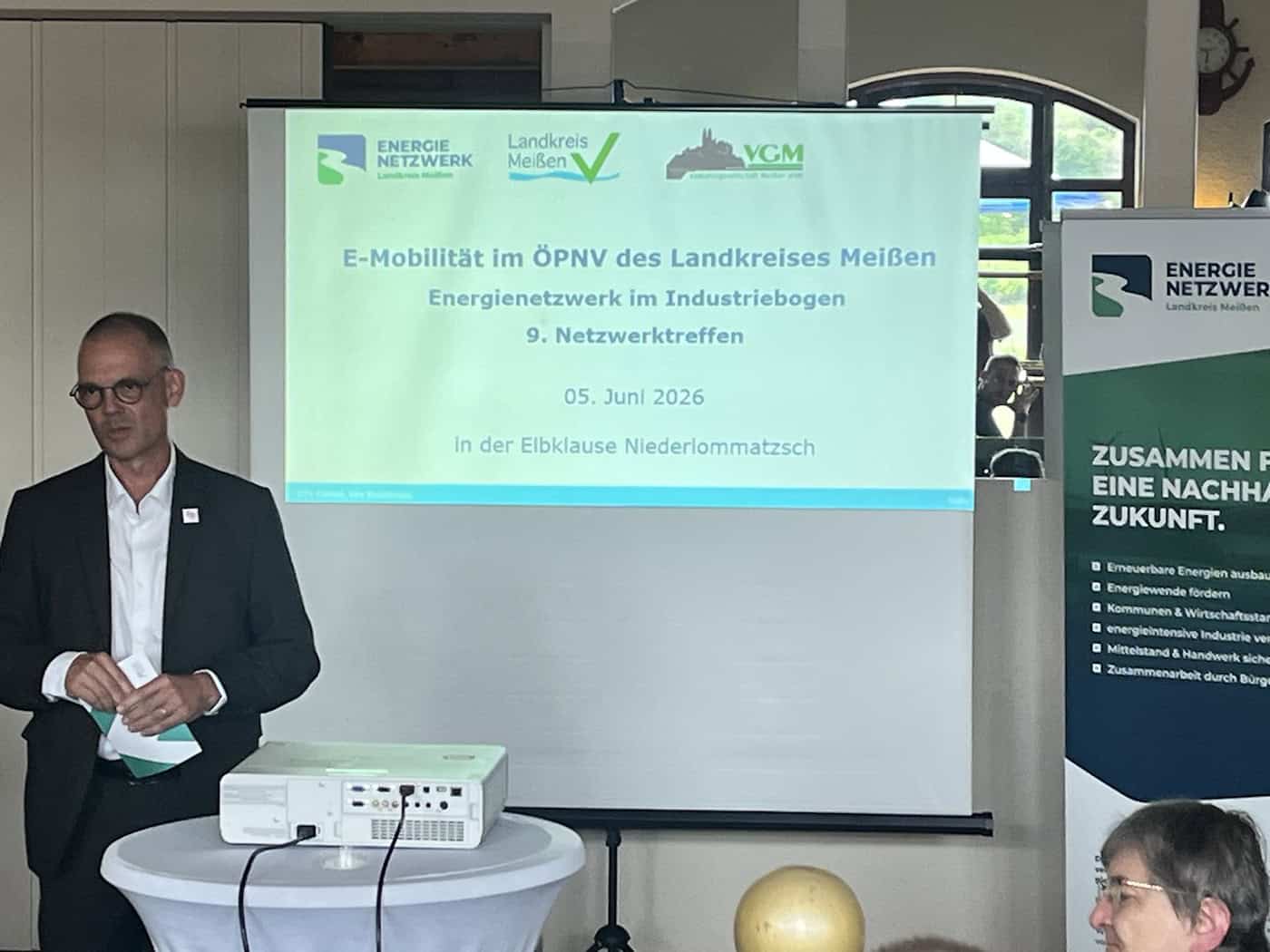 Foto: Mann im Anzug steht präsentierend vor einer Beamerprojektion mit dem Titel "E-Mobilität im ÖPNV des Landkreises Meißen"