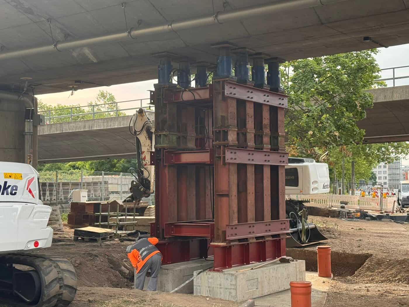 Eine Baustelle im Erdreich unter einer Betonbrücke. Eine massive, rotbraune Konstruktion aus schweren Stahlträgern stützt die Brückenunterseite ab. Im linken Vordergrund steht ein Bagger, während ein Bauarbeiter in orangefarbener Warnkleidung am Fundament arbeitet.