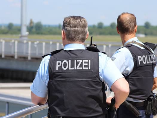 Zwei Polizisten von hinten. Sie tragen hellblaue Kurzarmhemden, schwarze Schutzwesten mit der weißen Aufschrift „POLIZEI“ und Einsatzgürtel mit Ausrüstung. Im Hintergrund ist ein verschwommenes Außengelände unter blauem Himmel zu sehen.