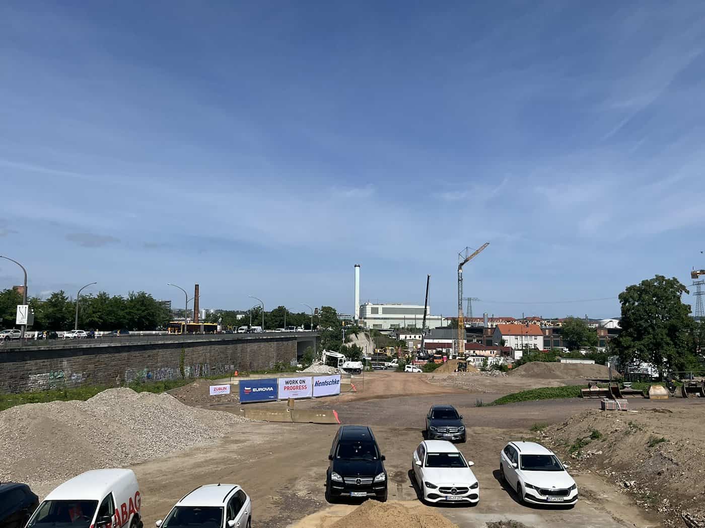 Blick von oben auf ein weitläufiges, sandiges Baustellengelände in Dresden. Im Vordergrund stehen mehrere Pkw und Transporter, im Hintergrund sind Bauzäune mit Firmenlogos von Eurovia und Züblin, ein großer gelber Baukran sowie ein hoher Industrieschornstein unter strahlend blauem Himmel zu sehen.