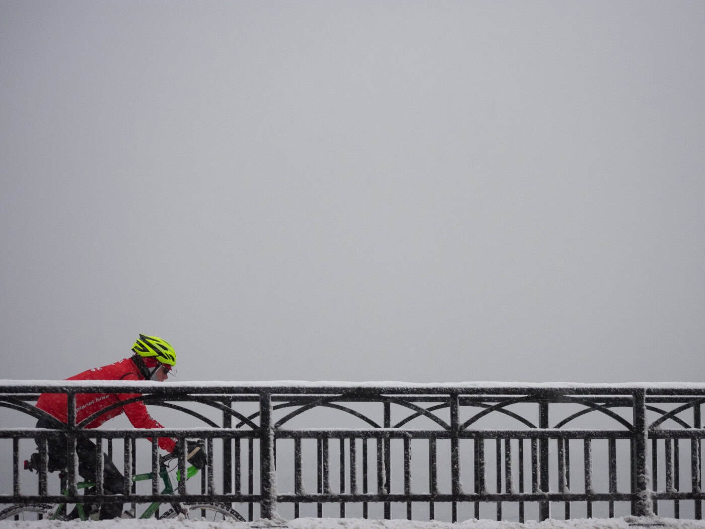 Eine Seitenansicht eines Radfahrers in einer leuchtend roten Sportjacke und einem neongelben Fahrradhelm, der von links nach rechts fährt. Er befindet sich hinter einem dunklen, gusseisernen Brückengeländer mit dekorativen Rundbögen, auf dem eine feine Schicht Schnee liegt.