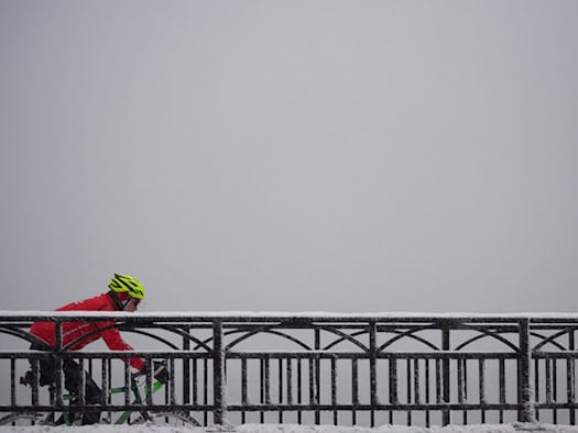 Eine Seitenansicht eines Radfahrers in einer leuchtend roten Sportjacke und einem neongelben Fahrradhelm, der von links nach rechts fährt. Er befindet sich hinter einem dunklen, gusseisernen Brückengeländer mit dekorativen Rundbögen, auf dem eine feine Schicht Schnee liegt.