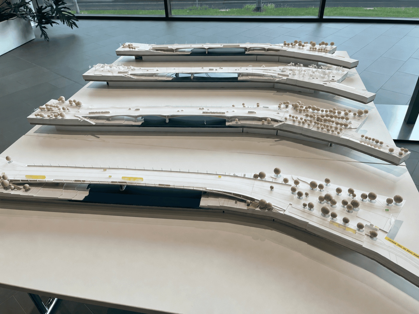 Ein Blick aus leicht erhöhter Perspektive auf vier filigrane, weiße Architekturmodelle einer Brücke, die hintereinander auf weißen Tischen in einem hellen Ausstellungsraum platziert sind. Jedes Modell stellt eine andere Brückenkonstruktion über einem schmalen, dunkelblauen Flusslauf dar. Die Uferber
