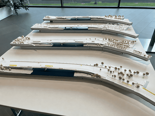 Ein Blick aus leicht erhöhter Perspektive auf vier filigrane, weiße Architekturmodelle einer Brücke, die hintereinander auf weißen Tischen in einem hellen Ausstellungsraum platziert sind. Jedes Modell stellt eine andere Brückenkonstruktion über einem schmalen, dunkelblauen Flusslauf dar. Die Uferber