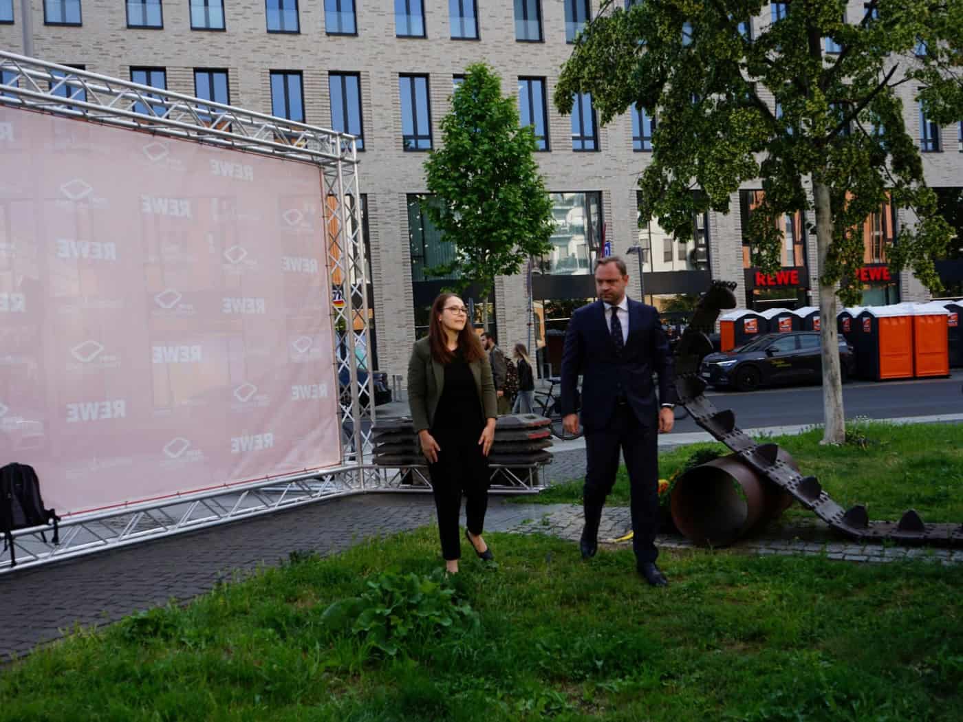 Eine Frau und ein Mann im Anzug stehen auf einer Wiese neben einem Denkmal aus einer Panzerkette. Links steht eine große, rosa Fotowand mit REWE-Logos vor einem modernen Gebäude im Hintergrund.