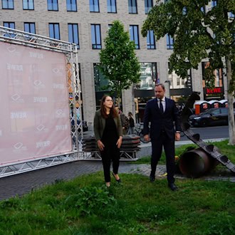Eine Frau und ein Mann im Anzug stehen auf einer Wiese neben einem Denkmal aus einer Panzerkette. Links steht eine große, rosa Fotowand mit REWE-Logos vor einem modernen Gebäude im Hintergrund.