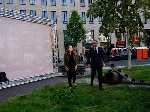 Eine Frau und ein Mann im Anzug stehen auf einer Wiese neben einem Denkmal aus einer Panzerkette. Links steht eine große, rosa Fotowand mit REWE-Logos vor einem modernen Gebäude im Hintergrund.