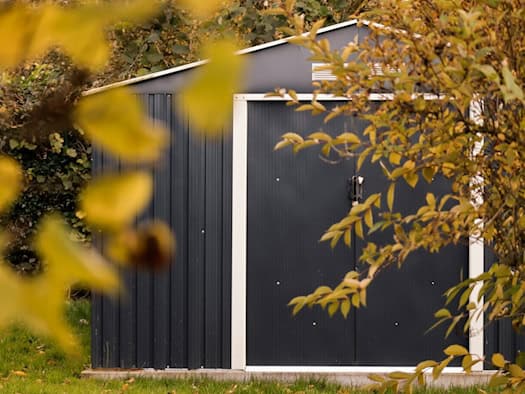 Dunkle Metallgarage im Garten, umgeben von herbstlichen Blättern und Sträuchern.