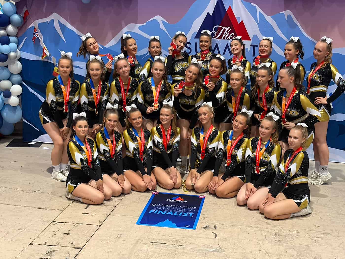 Das Dresdner Cheerleader-Team Allstars mit Medaillen als Finalist der European Summit 2026 Eine Gruppe junger Cheerleaderinnen kniet und steht lächelnd vor einer lila-blauen Bergkulissen-Fotowand. Sie tragen schwarz-goldene Outfits, rote Medaillen um den Hals und weiße Schleifen im Haar. Vor ihnen liegt ein blaues Finalisten-Schild.