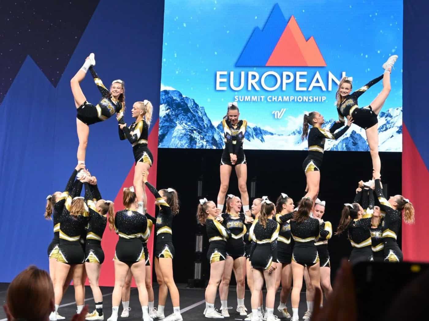 Cheerleading-Team in schwarz-goldenen Uniformen zeigt eine komplexe Hebefigur auf einer Bühne. Zwei äußere Athletinnen strecken ein Bein flexibel nach oben. Im Hintergrund zeigt eine LED-Wand das Logo „EUROPEAN SUMMIT CHAMPIONSHIP“.