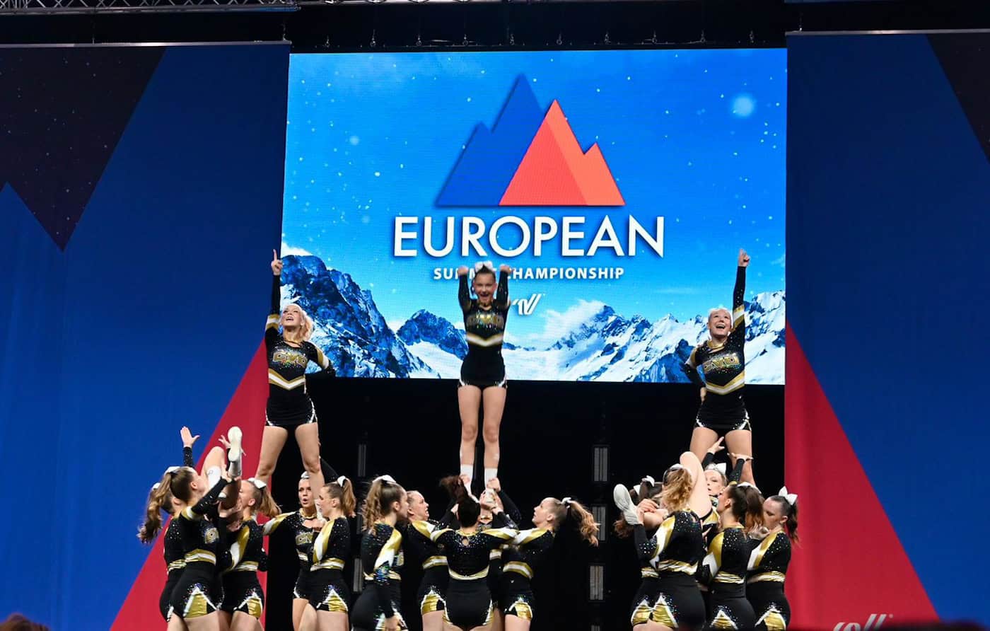 Cheerleader des CVD Dresden bei einer Hebefigur auf der Meisterschaftsbühne in Lissabon Drei Cheerleaderinnen werden von ihrem Team hoch in die Luft gestreckt und recken synchron den rechten Arm nach oben. Im Hintergrund leuchtet eine große blaue LED-Wand mit der Aufschrift „EUROPEAN SUMMIT CHAMPIONSHIP“.