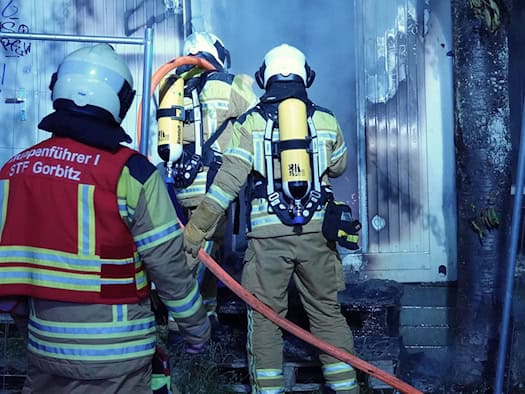Drei Feuerwehrleute im Einsatz vor einem verrußten Container. Ein Gruppenführer trägt eine rote Weste, zwei Atemschutzträger mit gelben Flaschen auf dem Rücken halten einen Löschschlauch.