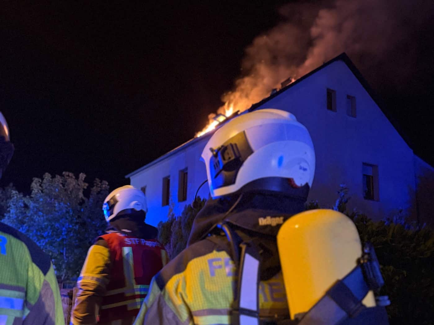 Feuerwehrleute mit Atemschutzausrüstung blicken im nächtlichen Blaulicht auf den lichterlohen Brand des Dachstuhls eines Wohnhauses in Dresden-Söbrigen.