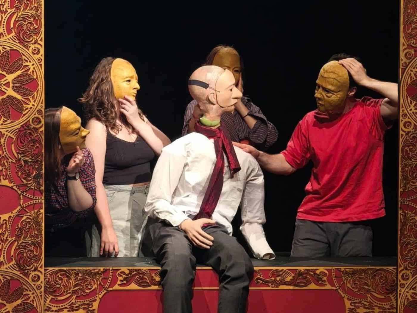Eine ausdruckslose Theaterpuppe im weißen Hemd sitzt auf einer rot-gold verzierten Bühne. Vier Schauspieler mit starren, gelben Masken umgeben sie vor schwarzem Hintergrund. Ein Darsteller im roten T-Shirt berührt die Schulter der Puppe.
