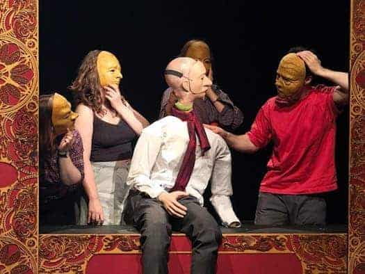 Eine ausdruckslose Theaterpuppe im weißen Hemd sitzt auf einer rot-gold verzierten Bühne. Vier Schauspieler mit starren, gelben Masken umgeben sie vor schwarzem Hintergrund. Ein Darsteller im roten T-Shirt berührt die Schulter der Puppe.