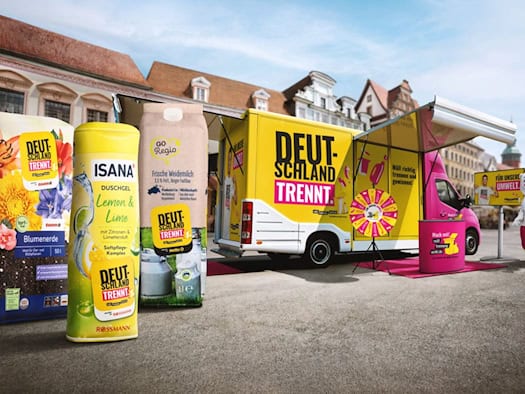 Ein großer gelb-weiß-roter Showtruck mit der Aufschrift „DEUTSCHLAND TRENNT.“ steht auf einem öffentlichen Platz. Davor sind drei riesige, aufgeblasene Nachbildungen von Produktverpackungen für Blumenerde, Duschgel und Weidemilch aufgestellt. 
