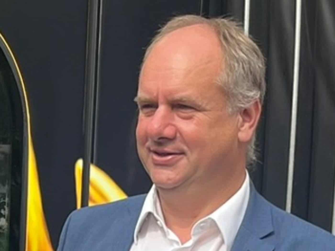 Porträtaufnahme von Dresdens Oberbürgermeister Dirk Hilbert im Dreiviertelprofil vor einem dunklen Hintergrund mit gelben Akzenten.  Er trägt ein blaues Sakko über einem offenen, weißen Hemd ohne Krawatte und blickt mit einem leichten Lächeln nach links.