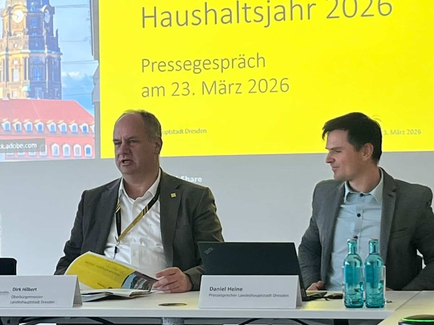 Drei Personen sitzen an einem Konferenztisch vor einer gelben Präsentationsfolie zum Haushaltsjahr 2026. In der Mitte spricht Dresdens Oberbürgermeister Dirk Hilbert.