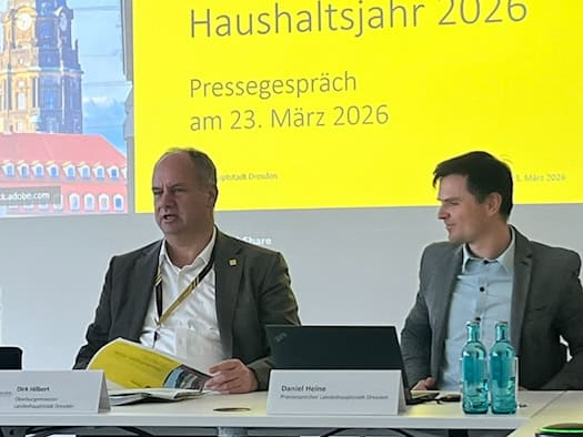 Drei Personen sitzen an einem Konferenztisch vor einer gelben Präsentationsfolie zum Haushaltsjahr 2026. In der Mitte spricht Dresdens Oberbürgermeister Dirk Hilbert.