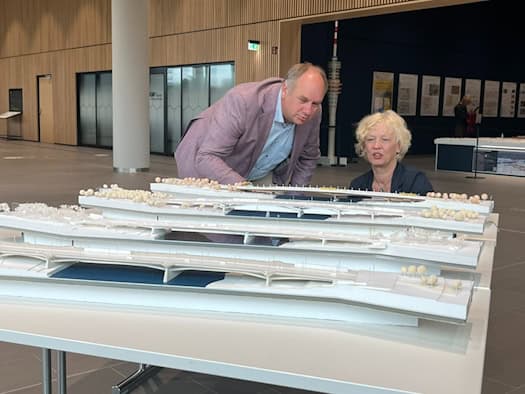 Ein Mann in einem dunklen Anzug und eine Frau stehen nebeneinander in einem hellen Ausstellungsraum des Stadtforums. Vor ihnen auf Tischen sind detaillierte, weiße Architekturmodelle von Brückenkonstruktionen aufgebaut, auf die beide im Gespräch blicken.