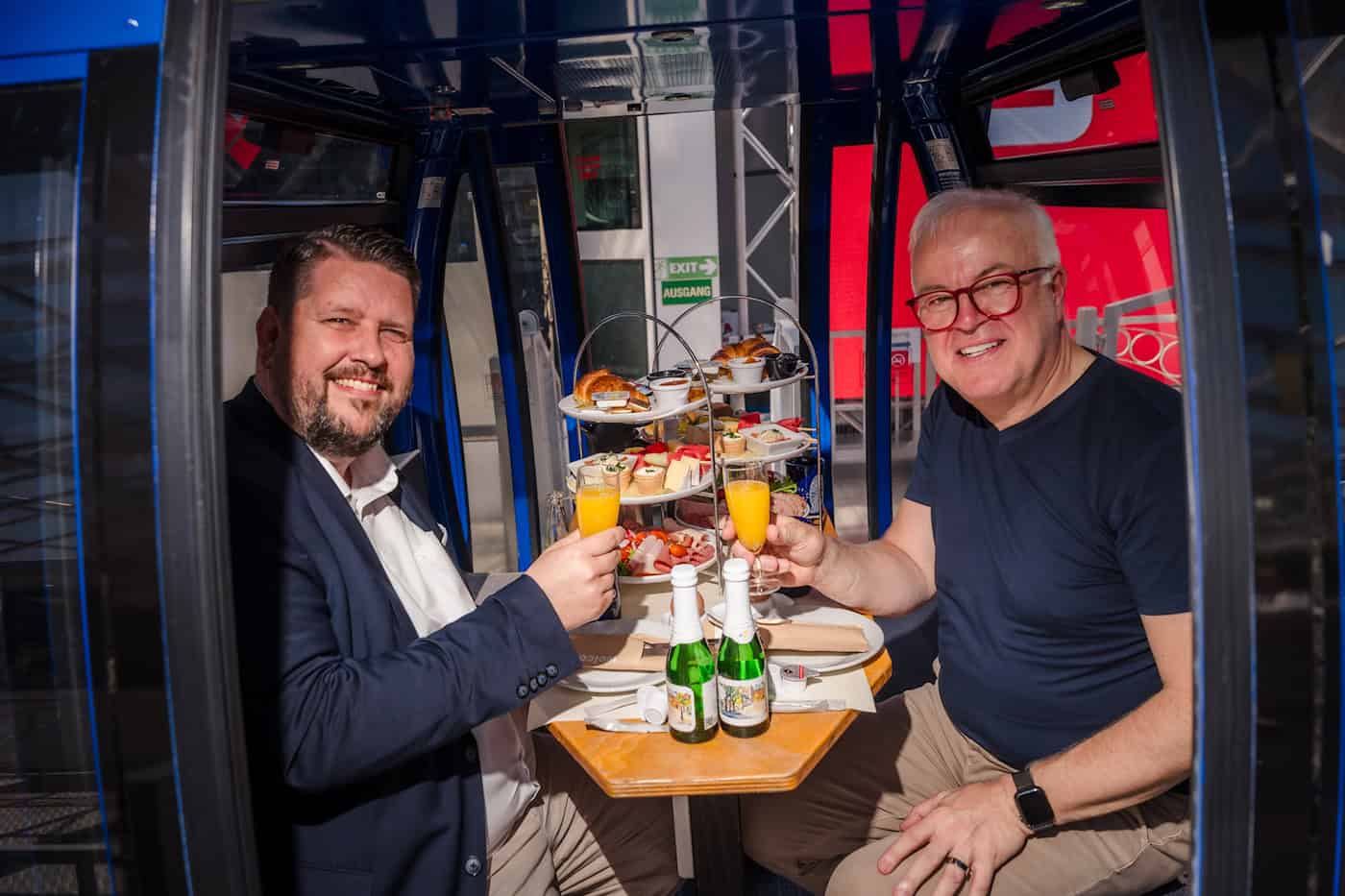 Henrik Oliver von Oehsen und Frank Schröder sitzen sich lächelnd in einer blauen Riesenrad-Gondel auf dem Postplatz gegenüber. Sie stoßen mit zwei Gläsern Orangensaft über einem gedeckten Frühstückstisch mit einer Etagere voller Snacks an.