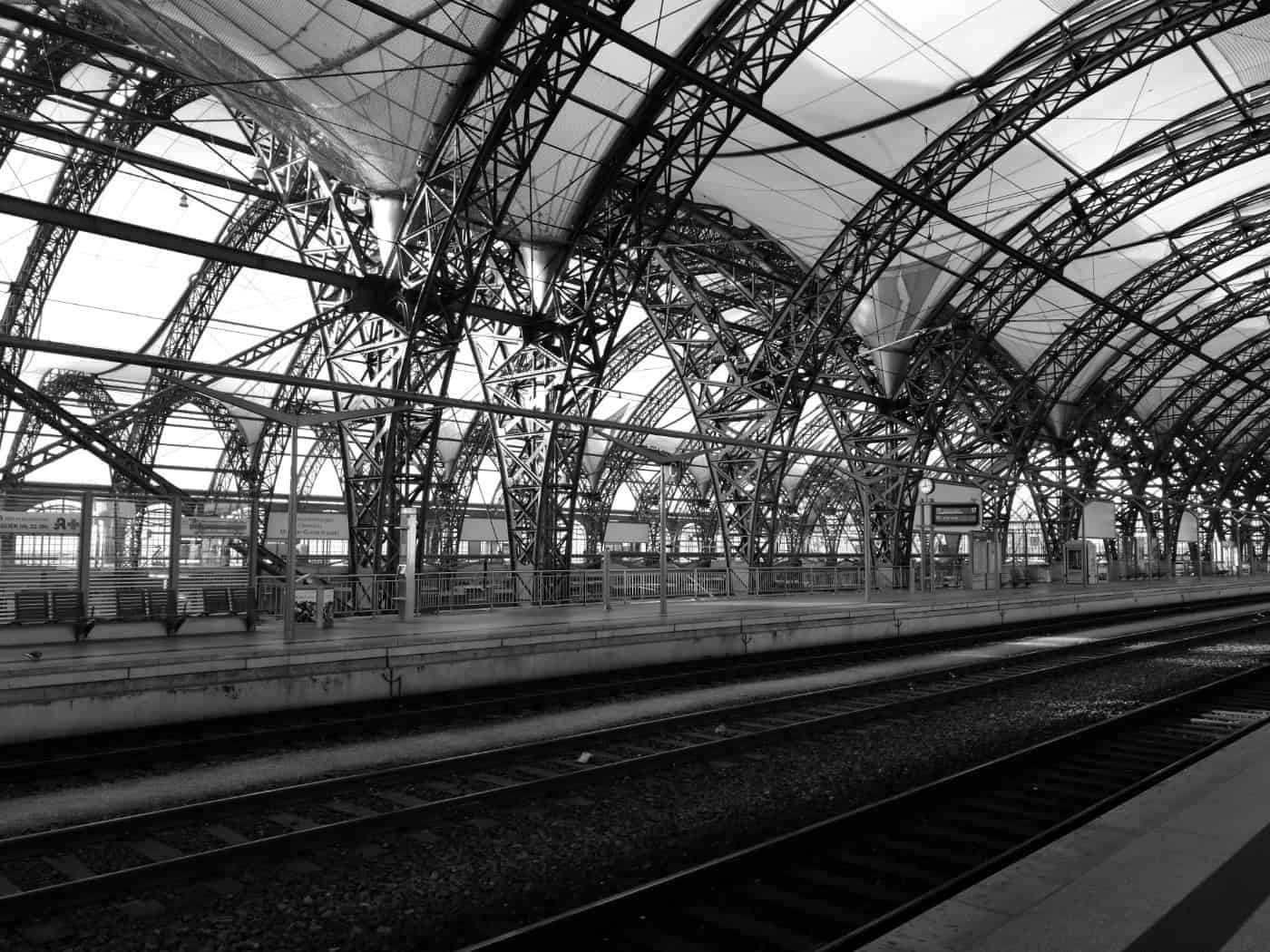 Innenansicht der weitläufigen, historischen Bahnhofshalle des Dresdner Hauptbahnhofs. Zu sehen ist ein moderner, gepflasterter Bahnsteig, an dem Züge halten. Die Konstruktion wird von einem großen, hellen Hallendach aus Stahl und Glas überspannt, Fahrgäste sind im Hintergrund zu erkennen.