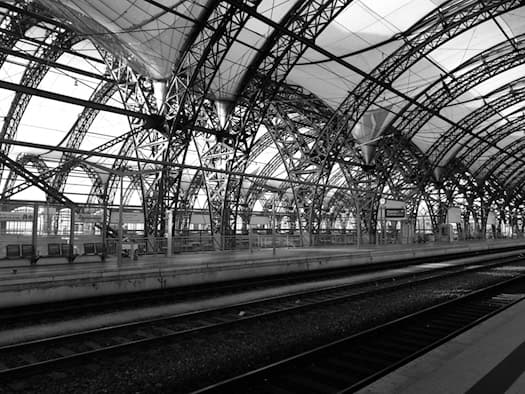 Innenansicht der weitläufigen, historischen Bahnhofshalle des Dresdner Hauptbahnhofs. Zu sehen ist ein moderner, gepflasterter Bahnsteig, an dem Züge halten. Die Konstruktion wird von einem großen, hellen Hallendach aus Stahl und Glas überspannt, Fahrgäste sind im Hintergrund zu erkennen.