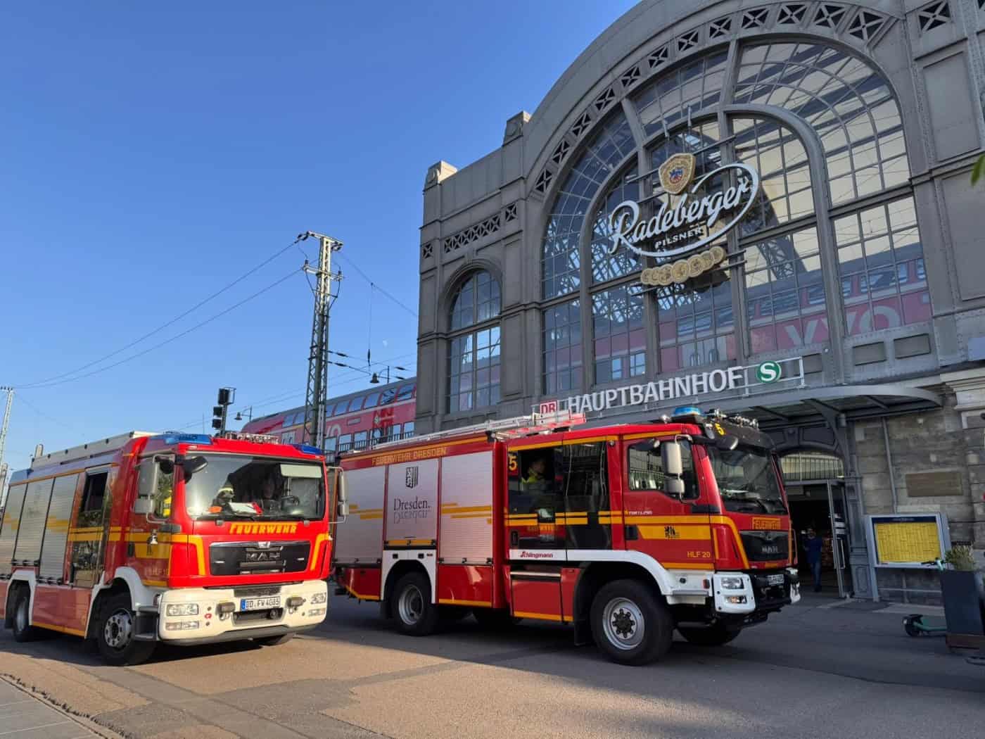 wei rote Einsatzfahrzeuge der Feuerwehr Dresden parken direkt vor dem historischen, gläsernen Haupteingang des Dresdner Hauptbahnhofs unter blauem Abendhimmel. Über dem Eingang sind der Schriftzug „Hauptbahnhof“ und ein großes Werbeschild zu sehen.