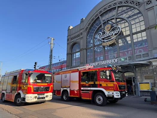 wei rote Einsatzfahrzeuge der Feuerwehr Dresden parken direkt vor dem historischen, gläsernen Haupteingang des Dresdner Hauptbahnhofs unter blauem Abendhimmel. Über dem Eingang sind der Schriftzug „Hauptbahnhof“ und ein großes Werbeschild zu sehen.