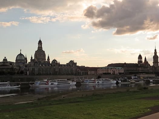 Panorama der historischen Altstadt von Dresden mit der Frauenkirche an der Elbe. Am Ufer liegen Raddampfer. Der Himmel ist von dichten, dunklen Wolken bedeckt, hinter denen die Sonne untergeht und ein kontrastreiches Licht wirft.