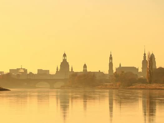 Die Silhouette der Dresdner Altstadt mit Frauenkirche spiegelt sich im ruhig fließenden Wasser der Elbe, während die gesamte Szenerie in ein warmes, goldenes Licht getaucht ist.