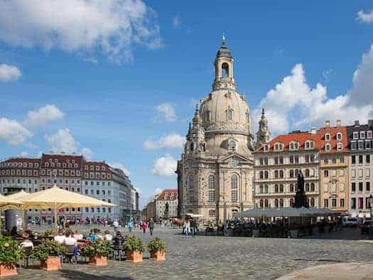 Die imposante barocke Frauenkirche steht zentral auf dem gepflasterten Neumarkt in Dresden unter blauem Himmel. Links sitzen Menschen im Außenbereich eines Cafés unter großen Sonnenschirmen, rechts säumen rekonstruierte Bürgerhäuser den Platz.