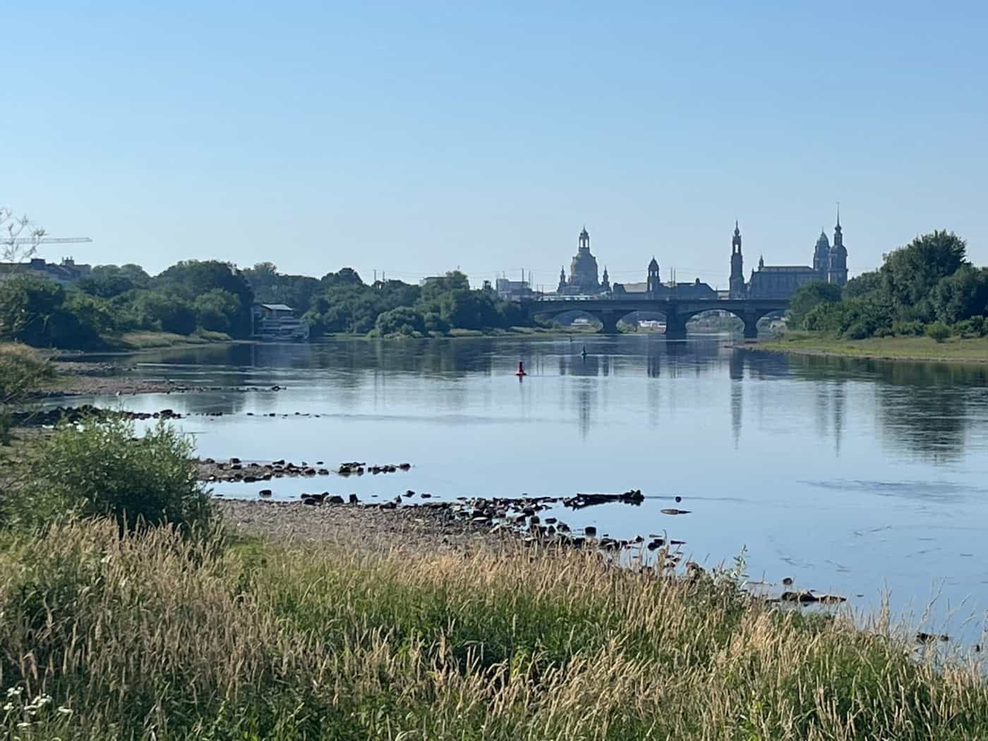 Blick über das extreme Niedrigwasser der Elbe auf die Silhouette der Dresdner Altstadt mit der Frauenkirche unter blauem Sommerhimmel. Im Vordergrund liegt eine breite, steinige Kiesbank komplett trocken.