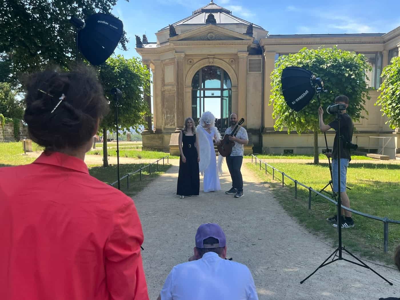 Ein Fotoshooting im sonnigen Park der Dresdner Elbschlösser vor einem historischen Pavillon-Gebäude. Im Hintergrund posieren eine Frau im schwarzen Abendkleid, ein Performer im weißen Engel-Kostüm mit Flügeln und Maske sowie ein Mann mit einer Akustikgitarre.