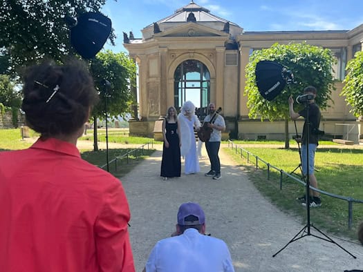 Ein Fotoshooting im sonnigen Park der Dresdner Elbschlösser vor einem historischen Pavillon-Gebäude. Im Hintergrund posieren eine Frau im schwarzen Abendkleid, ein Performer im weißen Engel-Kostüm mit Flügeln und Maske sowie ein Mann mit einer Akustikgitarre.