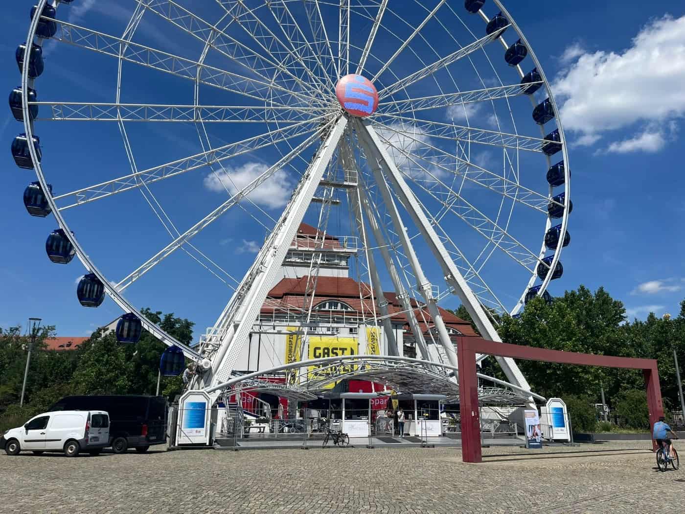 Das riesige weiße Riesenrad „Wheel of Vision“ mit blauen geschlossenen Gondeln steht auf dem gepflasterten Dresdner Postplatz. Im Hintergrund ist die markante Fassade des Schauspielhauses Dresden zu sehen.