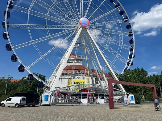 Das riesige weiße Riesenrad „Wheel of Vision“ mit blauen geschlossenen Gondeln steht auf dem gepflasterten Dresdner Postplatz. Im Hintergrund ist die markante Fassade des Schauspielhauses Dresden zu sehen.
