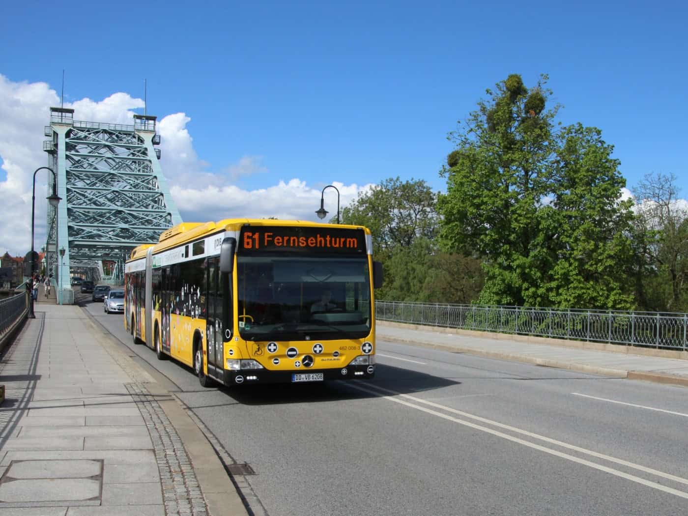 Ein gelb-schwarzer Gelenkbus der Dresdner Verkehrsbetriebe mit der Anzeige "61 Fernsehturm" fährt bei blauem Himmel über das stählerne Blaue Wunder in Dresden. Rechts befinden sich grüne Bäume.