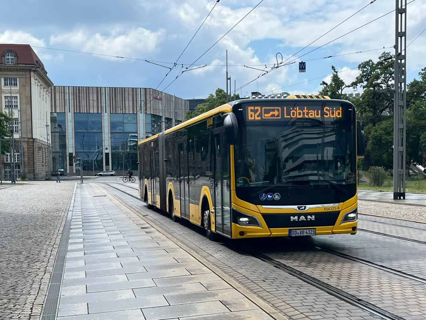 Ein moderner, gelb-schwarzer MAN-Gelenkbus der Dresdner Verkehrsbetriebe (DVB) fährt auf den im Pflaster eingelassenen Straßenbahnschienen einer Straße. Die digitale Linienanzeige an der Front des Busses zeigt „62 Löbtau Süd“. Im Hintergrund sind ein helles historisches Gebäude und ein moderner Neub