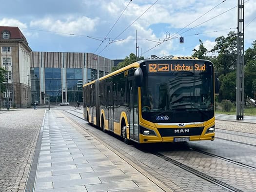 Ein moderner, gelb-schwarzer MAN-Gelenkbus der Dresdner Verkehrsbetriebe (DVB) fährt auf den im Pflaster eingelassenen Straßenbahnschienen einer Straße. Die digitale Linienanzeige an der Front des Busses zeigt „62 Löbtau Süd“. Im Hintergrund sind ein helles historisches Gebäude und ein moderner Neub