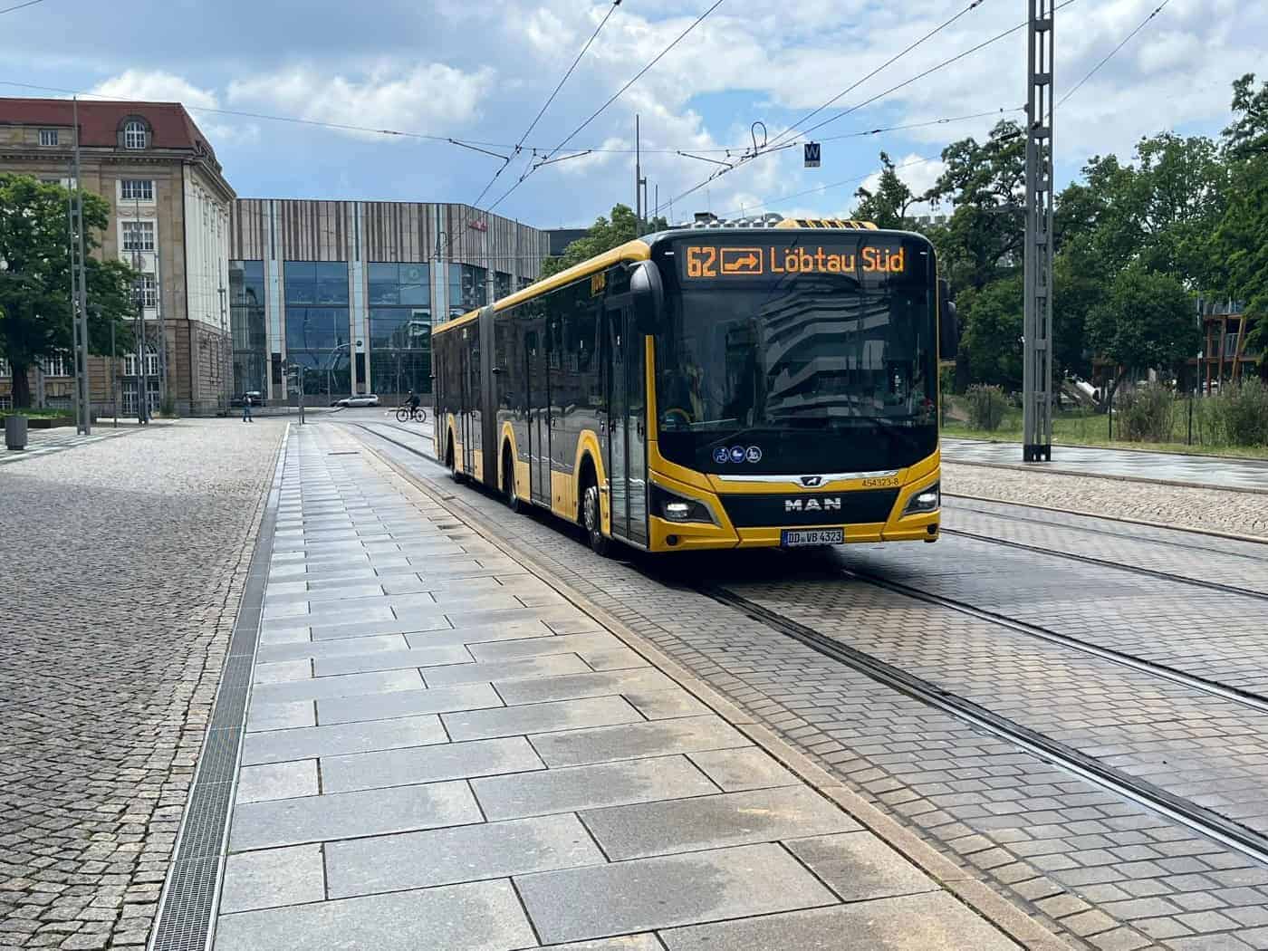 Ein gelb-schwarzer Gelenkbus der DVB mit der Anzeige „62 Löbtau Süd“ fährt auf einer gepflasterten Straße mit Schienen. Im Hintergrund sind moderne und historische Gebäude unter leicht bewölktem Himmel zu sehen.
